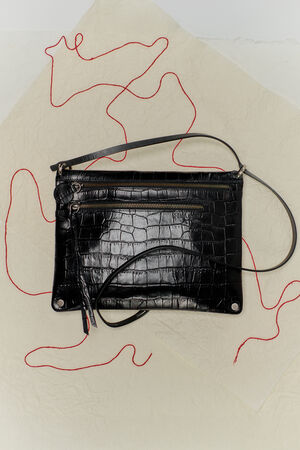 An INA KENT black crocodile-textured leather clutch with zippers, a detachable strap, and red thread loosely arranged around it on a textured beige surface.