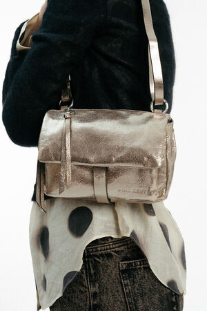 A person wearing a black jacket and printed blouse carries an INA KENT metallic gold crossbody bag with a tassel zipper detail.