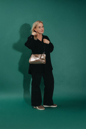 A woman stands against a green background, wearing a black outfit and brown shoes, holding an INA KENT metallic handbag over her shoulder.