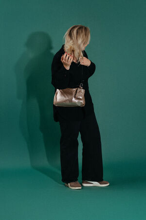 A person with blonde hair stands against a green background, wearing black clothing, tan shoes, and holding an INA KENT metallic shoulder bag. Their face is turned away from the camera.
