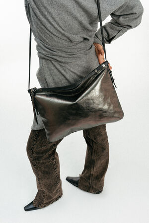 Eine Person, die ein graues Oberteil und eine braune Hose trägt, hält eine große INA KENT-Tasche aus schwarzem Metallic-Leder, die mit einem Reißverschluss verschlossen ist.