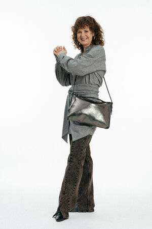 Frau mit lockigem Haar, die eine graue Strickjacke, eine gemusterte braune Hose und schwarze Schuhe trägt. Sie hält die Hände zusammen und trägt eine INA KENT-Metallic-Umhängetasche, die vor einem weißen Hintergrund steht.