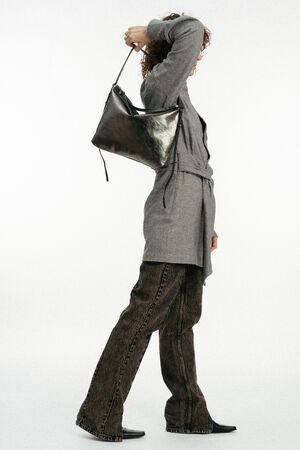 Eine Person in einem grauen Mantel mit Gürtel, einer braunen Hose und schwarzen Schuhen steht im Profil und trägt eine schwarze INA KENT-Ledertasche über der linken Schulter.
