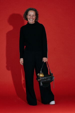 A person with short curly hair stands against a red background, smiling, wearing a black outfit and holding an INA KENT brown handbag with gold details.