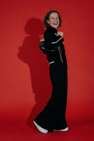 A person with curly hair stands against a red background, smiling and holding an INA KENT black handbag over their shoulder, wearing a black outfit and white shoes.