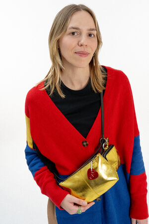 A woman wearing a red, blue, and yellow sweater and a black shirt poses with an INA KENT gold crossbody bag featuring a red keychain.