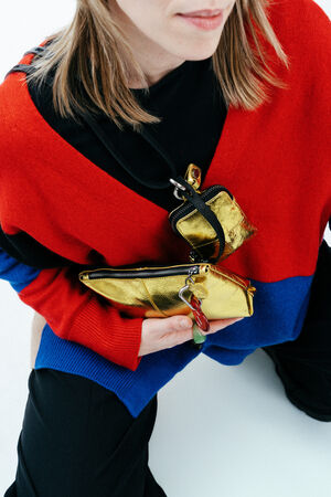 Person wearing a red and blue sweater, holding an INA KENT metallic gold clutch and a small matching pouch with keychain, photographed from above.