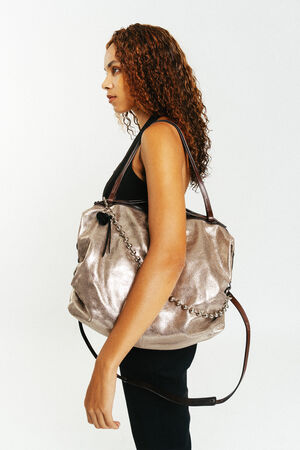 Frau mit lockigem Haar, die ein schwarzes ärmelloses Oberteil trägt und eine INA KENT Metallic-Tasche mit Kettendetails und braunen Trägern in der Hand hält, steht vor einem schlichten weißen Hintergrund.