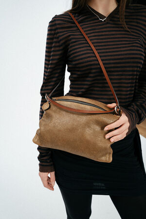 Eine Person, die ein braun-schwarz gestreiftes langärmeliges Hemd und einen schwarzen Rock trägt, hält eine braune INA KENT-Ledertasche mit Reißverschluss.
