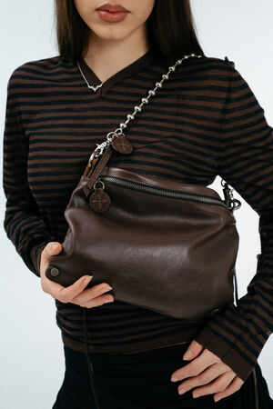 Eine Person, die ein braun-schwarz gestreiftes langärmeliges Hemd trägt, hält eine dunkelbraune INA KENT-Ledertasche mit einem silbernen Kettenriemen.