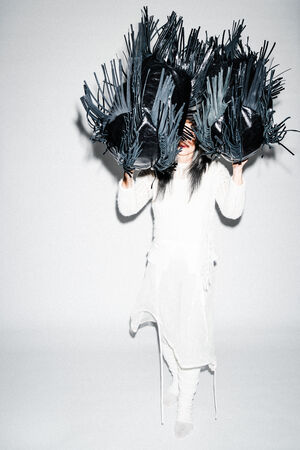 Eine weiß gekleidete Person steht vor einem hellen Hintergrund und hält zwei große schwarze INA KENT-Taschen, deren Fransen ihr Gesicht verdecken.