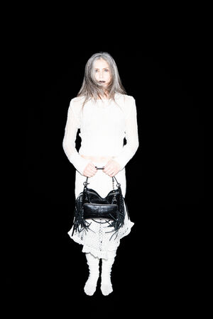 A woman in a white outfit stands against a black background, holding an INA KENT black fringed handbag in front of her with both hands.
