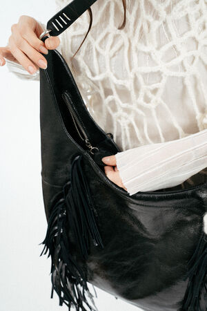 A person wearing a white textured sweater opens a large black INA KENT leather handbag with fringe detailing.