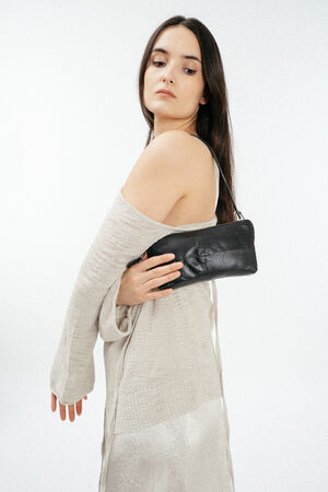 A woman in a cream knit sweater and white skirt holds an INA KENT black leather clutch under her arm against a plain white background.