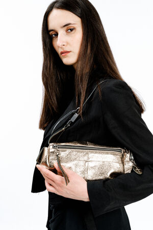 A woman with long dark hair, wearing a black blazer, holds an INA KENT metallic gold handbag under her arm against a white background.