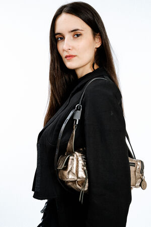 Woman with long dark hair wearing a black jacket and carrying an INA KENT metallic handbag over her shoulder, standing against a plain white background.