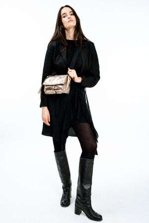 A woman in a black dress, black coat, and black knee-high boots stands facing forward, holding an INA KENT metallic shoulder bag against a plain white background.