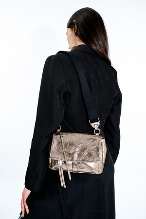 A person with long dark hair, wearing a black coat, stands facing away, showcasing an INA KENT metallic gold crossbody bag with a wide black strap.