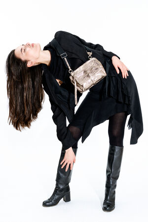 Woman in a black outfit and knee-high boots poses bending backward with an INA KENT metallic crossbody bag against a white background.