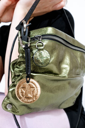 A person holds a metallic green INA KENT bag with a zipper and two round metallic keychains, one silver and one bronze, attached to the strap.