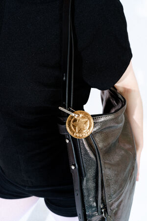 Person carrying an INA KENT metallic leather bag over their shoulder, with a large gold coin-shaped keychain attached to the bag's strap.