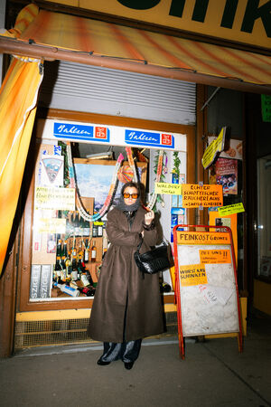 Person in a long brown coat and sunglasses stands in front of a small shop decorated with colorful signs, bottles, and sausage garlands, holding a cigarette and an elegant black INA KENT handbag.