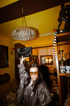 Eine Frau mit Sonnenbrille und gemustertem Mantel steht in einem Raum und hält eine schwarze INA KENT-Handtasche hoch. Der Raum ist mit einem Kronleuchter, Regalen, Kunst und verschiedenen Gegenständen dekoriert.