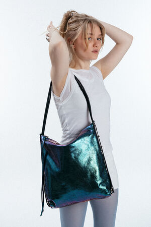 Eine Frau in einem weißen T-Shirt posiert mit den Händen in den Haaren und präsentiert eine große, schillernde INA KENT-Umhängetasche mit schwarzen Trägern vor einem schlichten weißen Hintergrund.