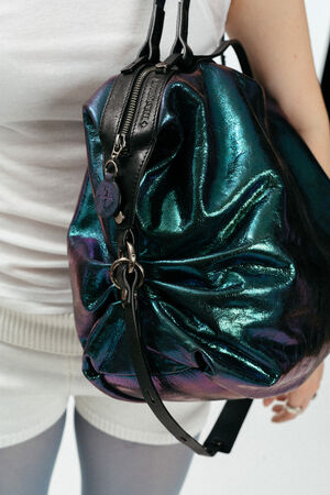 Eine Person, die ein weißes Oberteil und kurze Hosen trägt, hat eine große, glänzende, grün und blau schillernde INA KENT-Tasche mit schwarzen Trägern und Beschlägen bei sich.