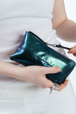 Person mit einer glänzenden, metallisch blauen INA KENT-Clutch-Tasche mit Reißverschluss und einem Schlüssel mit einem herzförmigen Schlüsselanhänger, bekleidet mit einem weißen ärmellosen Oberteil und einer weißen Hose.