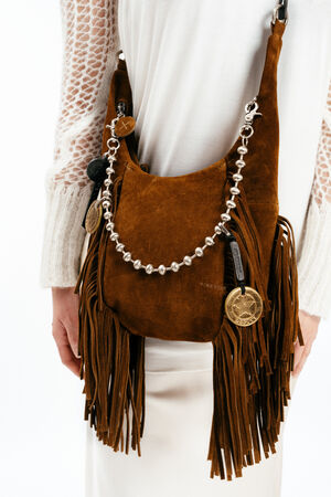 A person wearing a white outfit carries an INA KENT brown suede fringe shoulder bag with metallic and round embellishments and medallion charms.