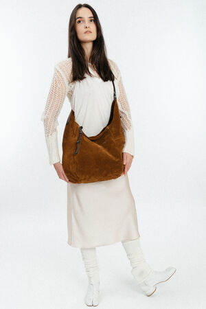 Woman standing against a plain background, wearing a cream-colored knit top, ivory skirt, white boots, and leg warmers, holding a large brown INA KENT suede shoulder bag.