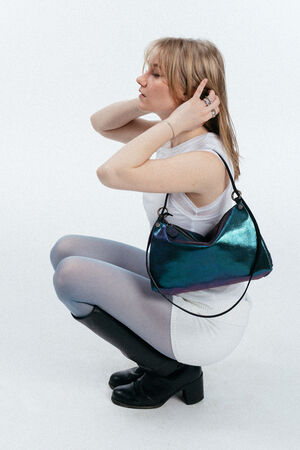 Eine Person mit hellem Haar hockt, trägt ein weißes Oberteil, einen weißen Rock, eine durchsichtige Strumpfhose, schwarze Stiefel und hält eine glänzende blau-grüne INA KENT-Handtasche.