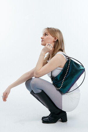 Eine Frau in weißem Outfit und schwarzen Stiefeln hockt, den Blick zur Seite gerichtet, mit einer metallisch schillernden Handtasche von INA KENT über der Schulter vor einem schlichten weißen Hintergrund.