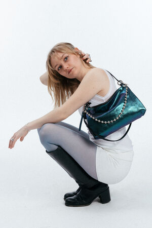 Eine Frau in weißem Outfit und schwarzen Stiefeln hockt sich hin und blickt in die Kamera, während sie eine metallicblaue INA KENT-Handtasche mit einem Kettenriemen über der Schulter trägt.