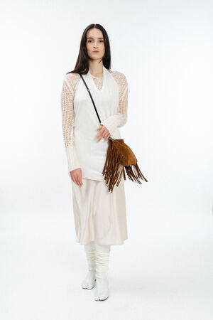 A woman stands against a white background wearing a long white dress with mesh sleeves, white boots, and an INA KENT brown fringed crossbody bag.