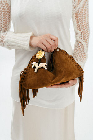 Person holding an INA KENT brown suede handbag with fringe details and a gold horse-shaped charm, wearing a white long-sleeve top with netted sleeves.