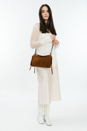 A woman wearing a white long-sleeve top, cream skirt, leg warmers, and white shoes stands holding an INA KENT brown suede shoulder bag against a plain white background.