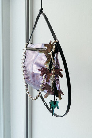 Eine lavendelfarbene INA KENT-Handtasche mit Perlenriemen, verziert mit mehreren bunten Schlüsselanhängern in Tierform, hängt an einem schwarzen Riemen vor einer weißen Wand.