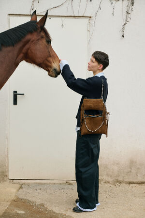 A person standing in front of a closed white door pets a brown horse. Wearing loose dark pants, a black jacket, and an INA KENT brown bag over their shoulder, they exude effortless style.