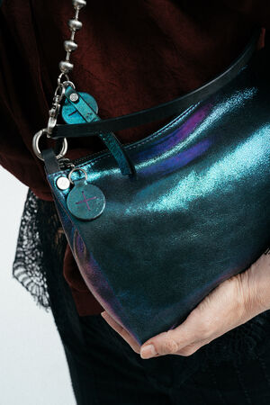 Eine Person hält eine metallisch schillernde Handtasche von INA KENT mit einem Kettenriemen in der Hand. Sie trägt ein dunkelrotes Oberteil und eine dunkle Hose.