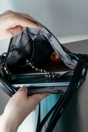 A person opens an INA KENT black handbag, showing the contents inside, including a silver beaded keychain, an apple, and a laptop.