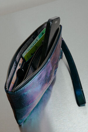 A small, iridescent INA KENT zippered pouch containing a smartphone, cards, cash, and a black item rests on a metallic surface.