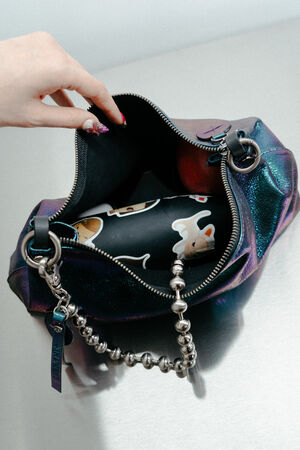 A hand opens a zipped iridescent INA KENT purse with a metallic ball chain strap, revealing a black water bottle decorated with animal stickers and an apple inside.
