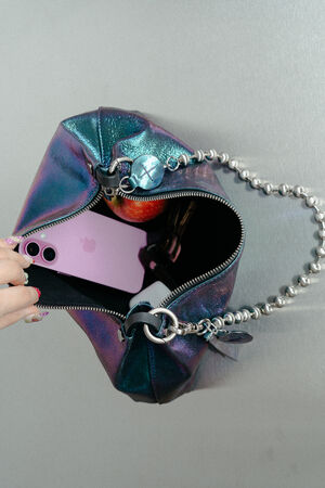 A hand opens an INA KENT metallic purple handbag with a silver chain strap, revealing a pink smartphone and keys inside.