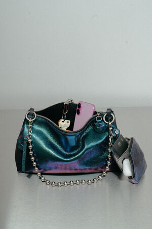 A metallic, iridescent INA KENT handbag with a beaded chain strap is open, revealing a pink smartphone, keys, and a small attached pouch containing wireless earbuds.