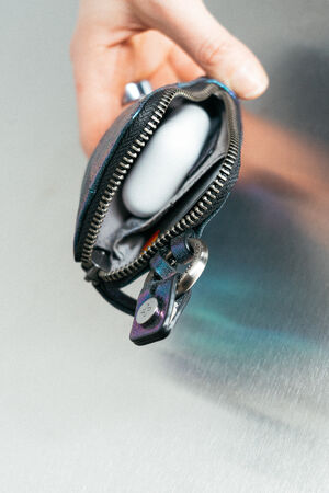 A hand holds an open INA KENT zippered pouch containing a white wireless earbud case against a metallic background.