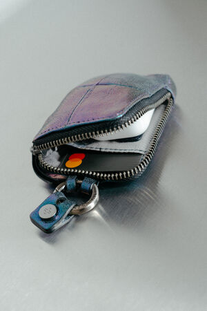 A small INA KENT zippered pouch lies on a metallic surface, partially open to reveal a credit card and a set of white wireless earbuds inside.