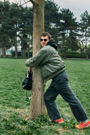 A person wearing sunglasses, a green jacket, and orange shoes leans against a tree in a grassy park, holding a black INA KENT bag.