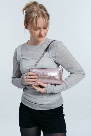 A woman wearing a light gray long-sleeve top and black shorts holds an INA KENT metallic pink handbag with a chain strap, looking down at the bag.